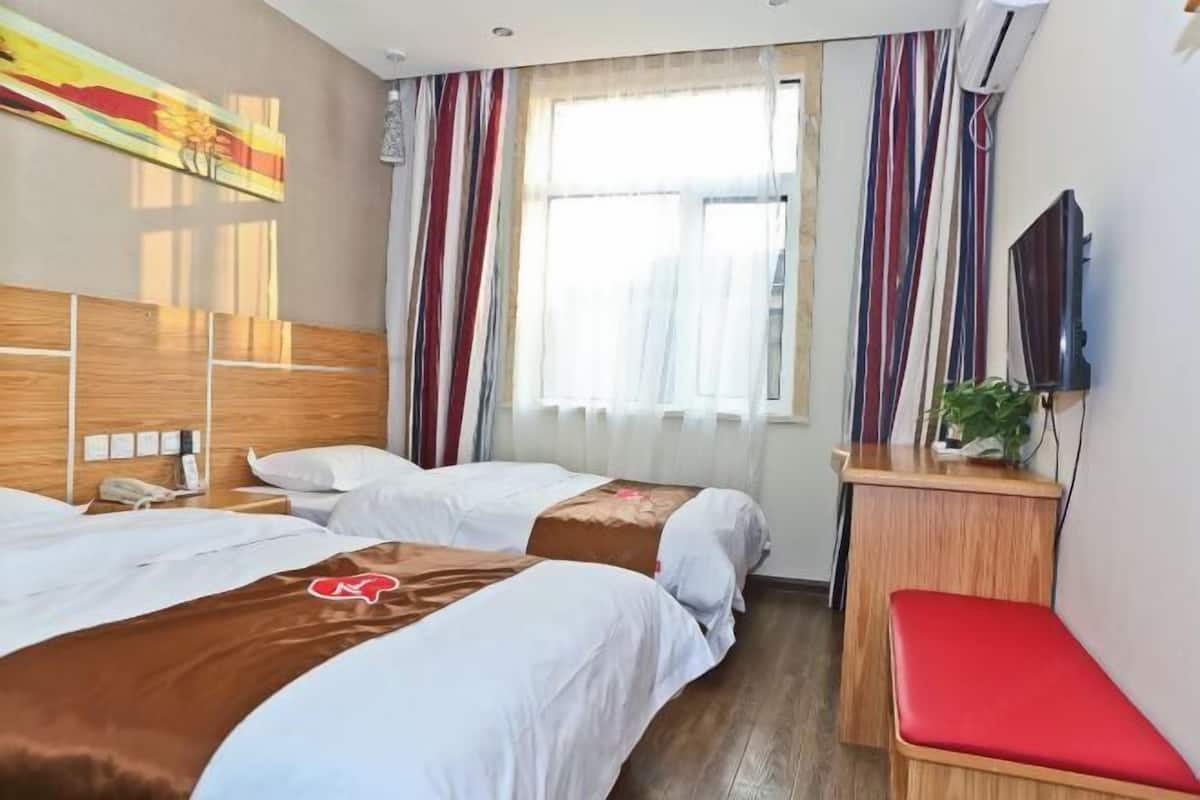 A hotel room with two beds, a desk with a red chair, a TV, and a window with curtains.