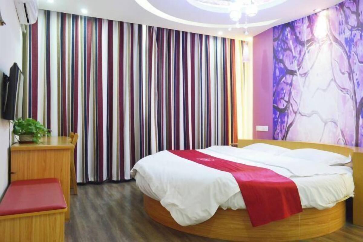 A hotel room with a round bed, a wooden desk, a red bench, a television, and a curtain with vertical stripes.