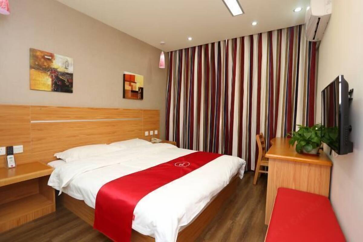 A hotel room with a wooden headboard, a bed with a red and white bedspread, a desk with a chair, a television, and a striped curtain.
