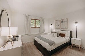 Une chambre à coucher comprenant un lit, des tables de chevet, une lampe, une fenêtre avec des rideaux et des œuvres d’art murales.