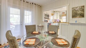 A dining area with a glass table surrounded by wicker chairs, a kitchen in the background, and a view through a window with sheer curtains. - Unwind Fireside & Walk to Dtwn! Tyron Retreat (Tryon)