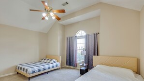 Une chambre avec deux lits, un ventilateur de plafond, une fenêtre avec des rideaux et une petite table avec une lampe.