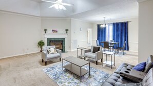 Un salon avec une cheminée, deux fauteuils, un canapé, une table basse et un coin repas agrémenté de rideaux bleus.
