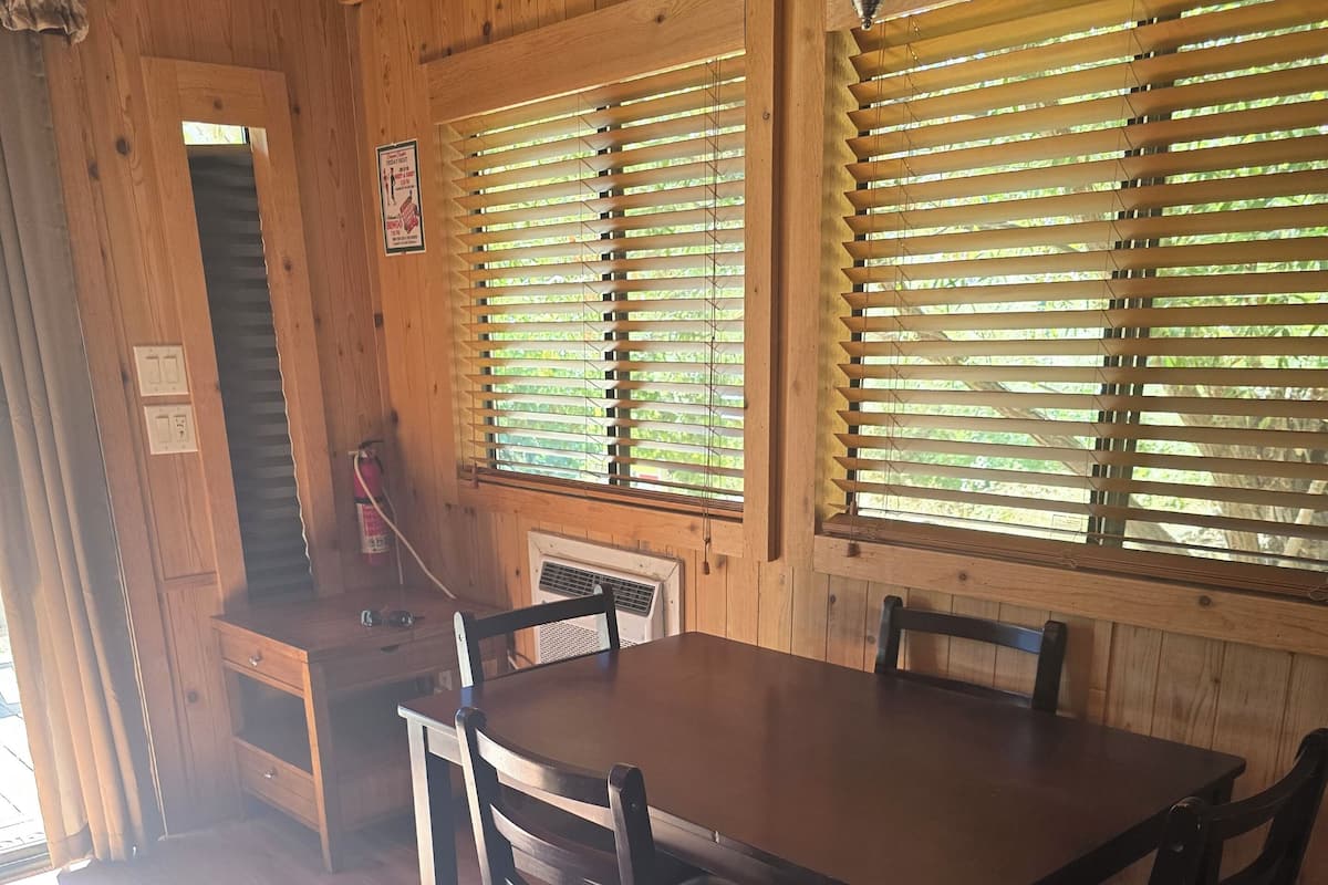 A wooden dining area with a table and chairs, large windows with blinds, and a ceiling fan.
