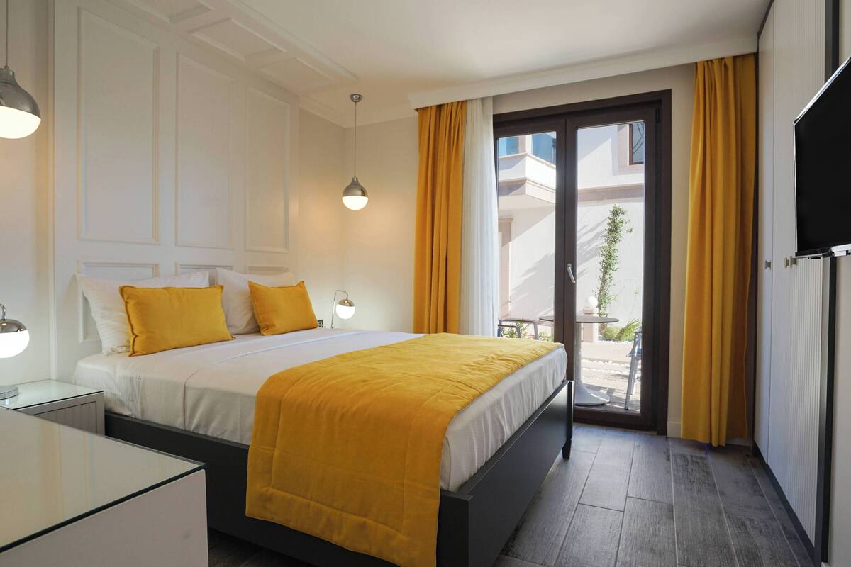 A hotel room with a bed, bedside tables, a TV, and a balcony with yellow curtains.