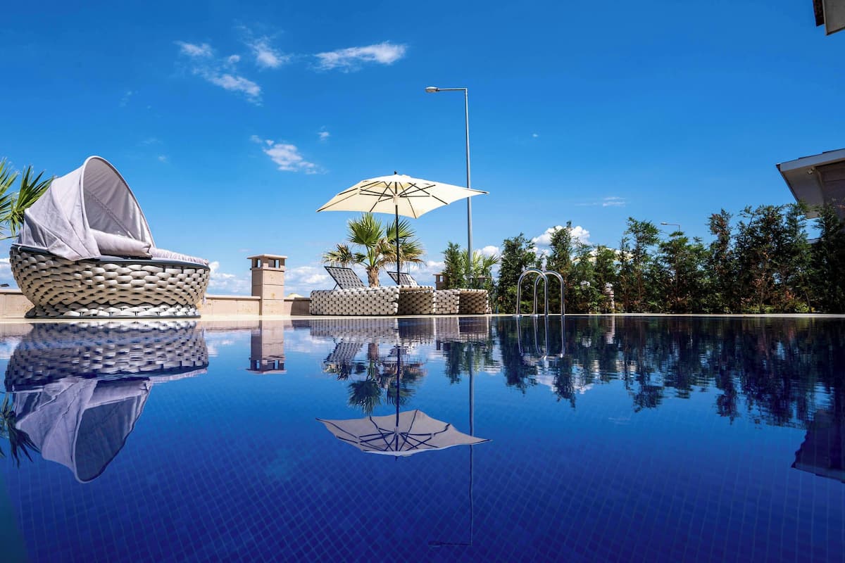 A pool area with lounge chairs and umbrellas, surrounded by palm trees and a clear sky.