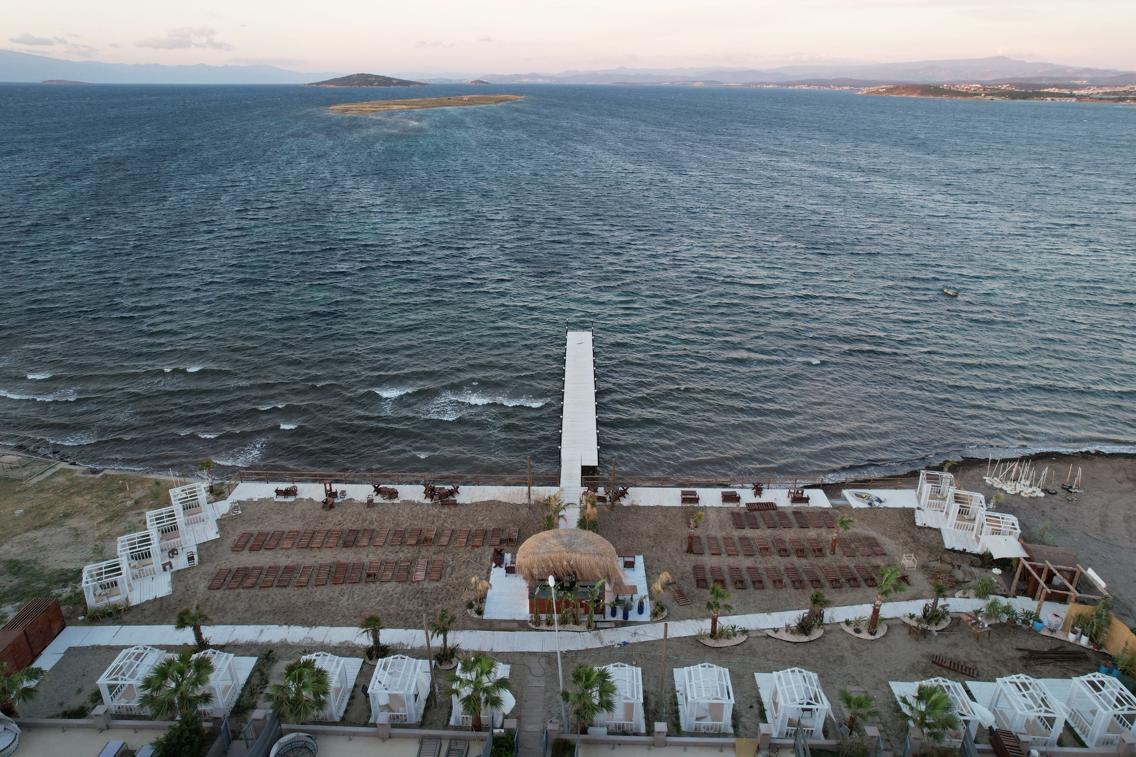 Vue aérienne d’un complexe hôtelier en bord de mer, avec un bâtiment central entouré de rangées de sièges et de palmiers.