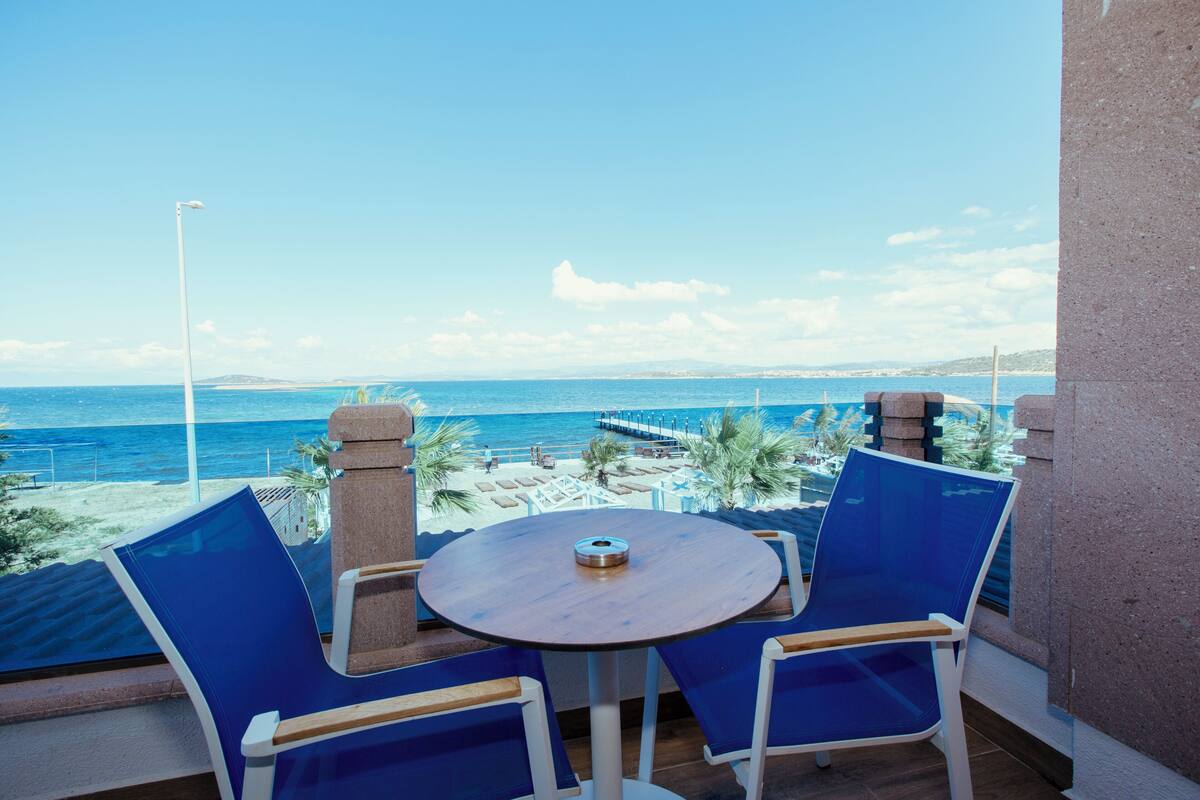 A balcony with a round table and blue chairs, overlooking a beach and the sea.