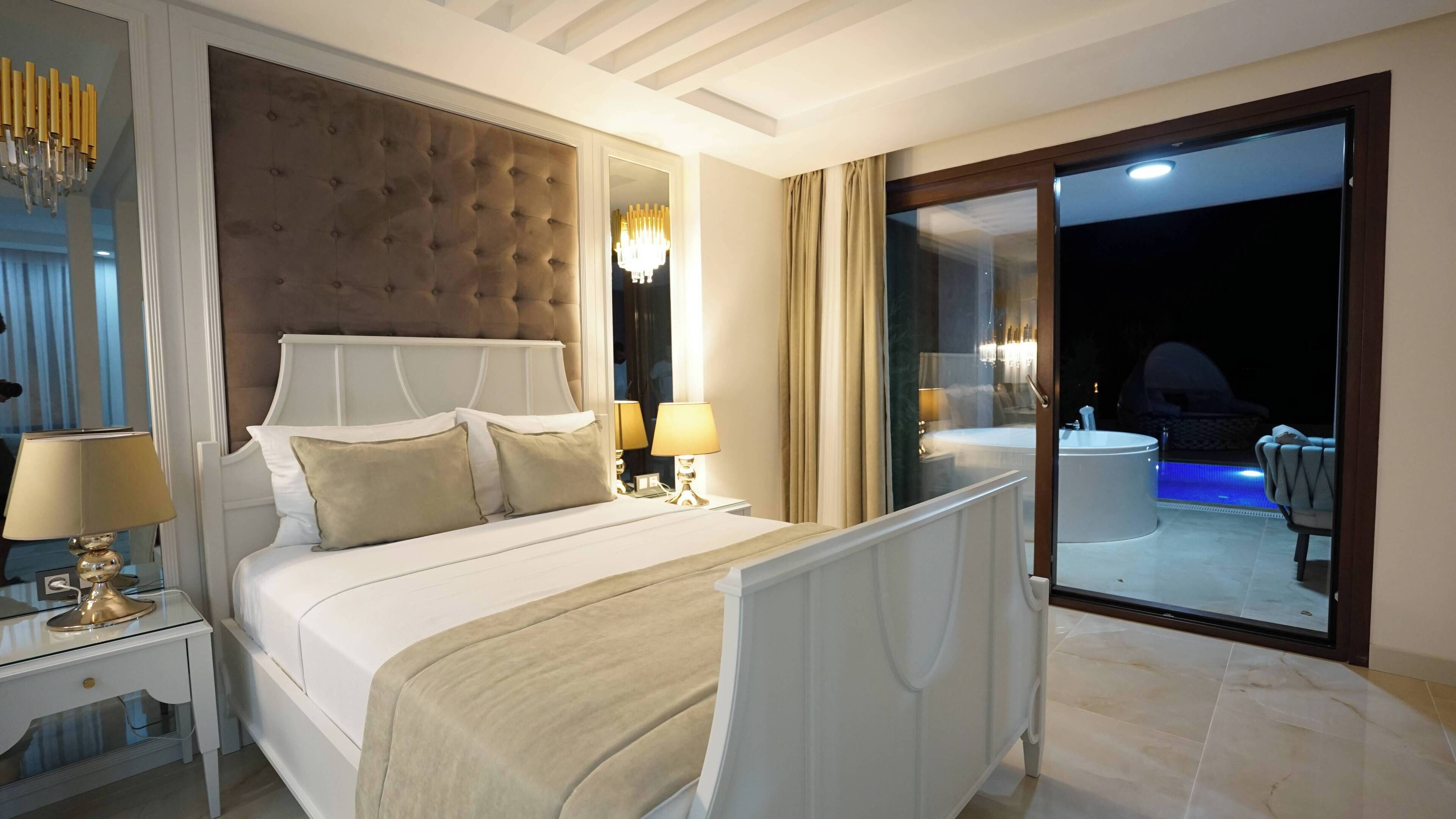 Ein geräumiges Schlafzimmer mit einem großen Bett, Nachttischlampen, einem gepolsterten Kopfteil und Blick auf einen Poolbereich.