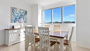 A dining area with a wooden table and white chairs, a white cabinet, and a large window with blinds. - Boronia Beach House One - 650m to headland (Sawtell)