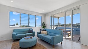 A modern living room with two blue sofas, a small ottoman, and a large window offering a view of the beach and ocean. - Boronia Beach House One - 650m to headland (Sawtell)
