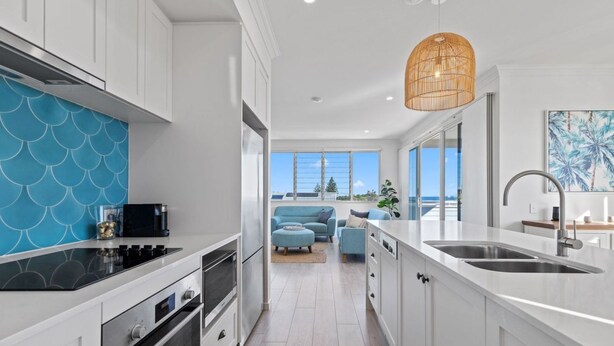A modern kitchen with white cabinetry, a blue backsplash, and a large window offering a view of the outdoors. - Boronia Beach House One - 650m to headland (Sawtell)