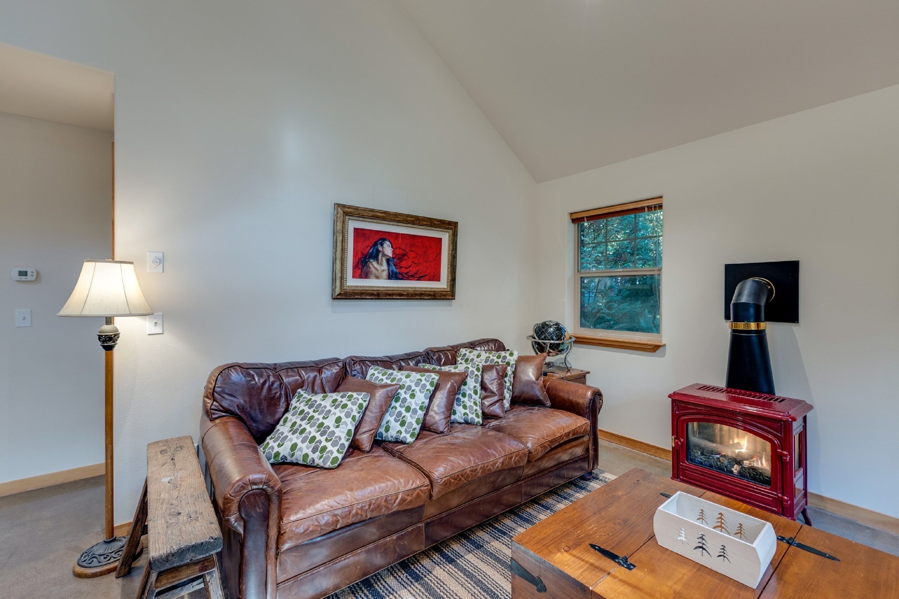 A living room with a brown leather sofa, patterned pillows, a wooden coffee table, a red wood-burning stove, a floor lamp, and a framed painting on the wall.