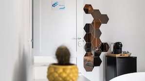 A modern hotel room with a white door, a sign with a logo, a geometric wall decoration, a small cactus, a black coffee machine, and a wooden side table.