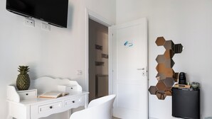 A modern, well-lit room with a white desk, a white chair, a flat-screen TV mounted on the wall, and a door with a hexagonal mirror decoration.
