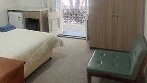 A hotel room with a bed, a desk with a computer, a television, a wardrobe, and a balcony with a view of trees. - Woolpack Hotel (Parramatta)