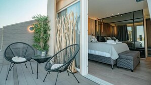 A modern bedroom with a large bed, a small seating area with chairs, and a potted plant.