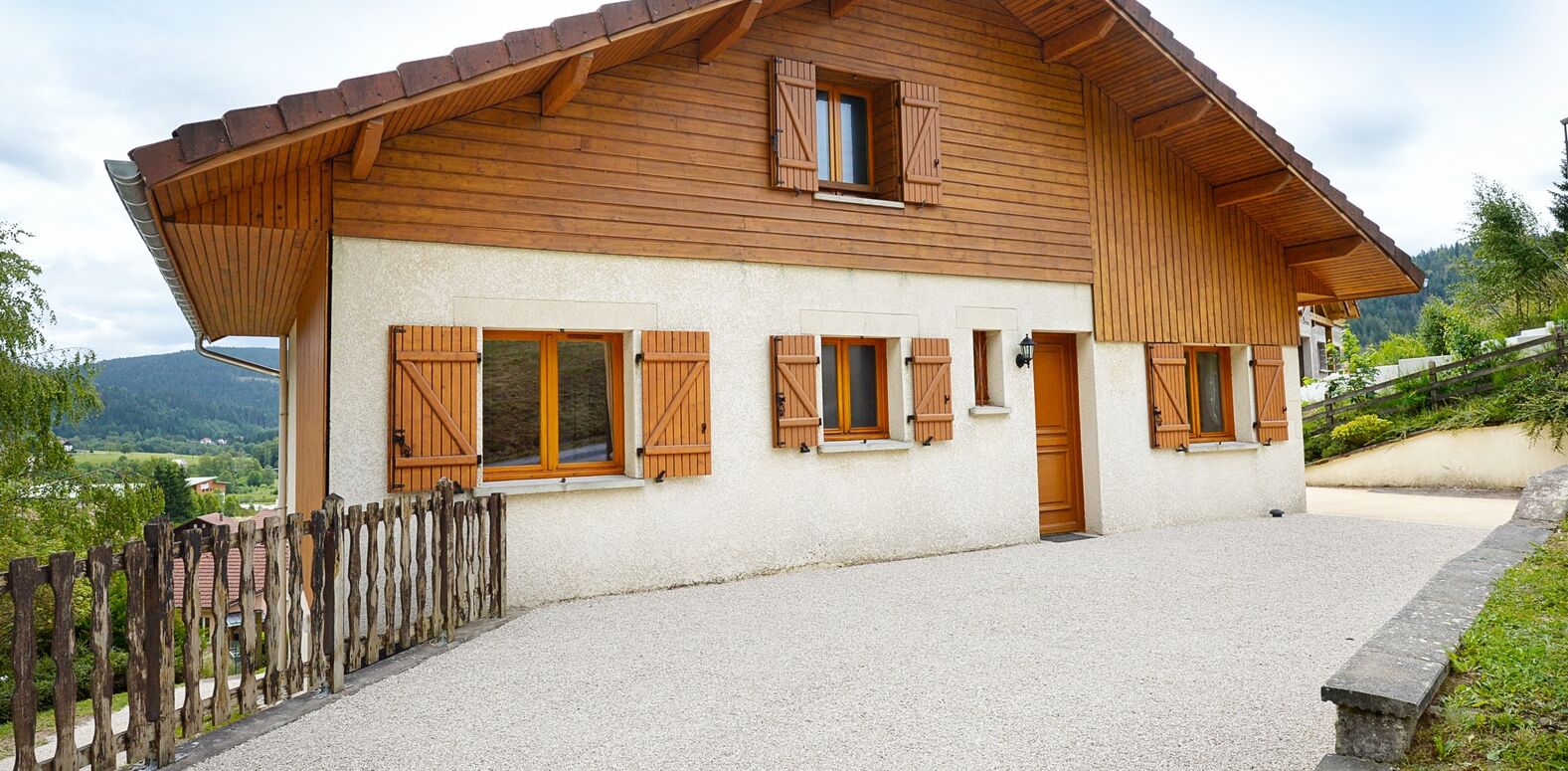 A wooden chalet with a sloped roof, multiple windows with shutters, and a gravel driveway.
