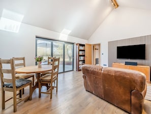 A modern living room with a wooden dining table, chairs, a brown leather sofa, a flat-screen TV, and a bookshelf.