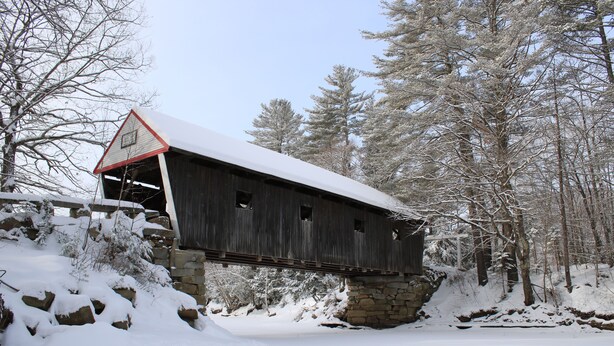 A wooden covered bridge with a triangular roof, supported by stone pillars, in a snowy forest setting.