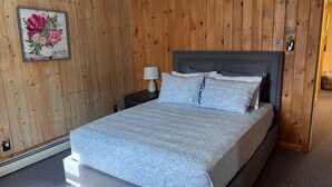 A bedroom with a wooden paneled wall, a bed with a floral-patterned bedspread, two matching pillows, a nightstand with a lamp, and a framed floral artwork above the bed.