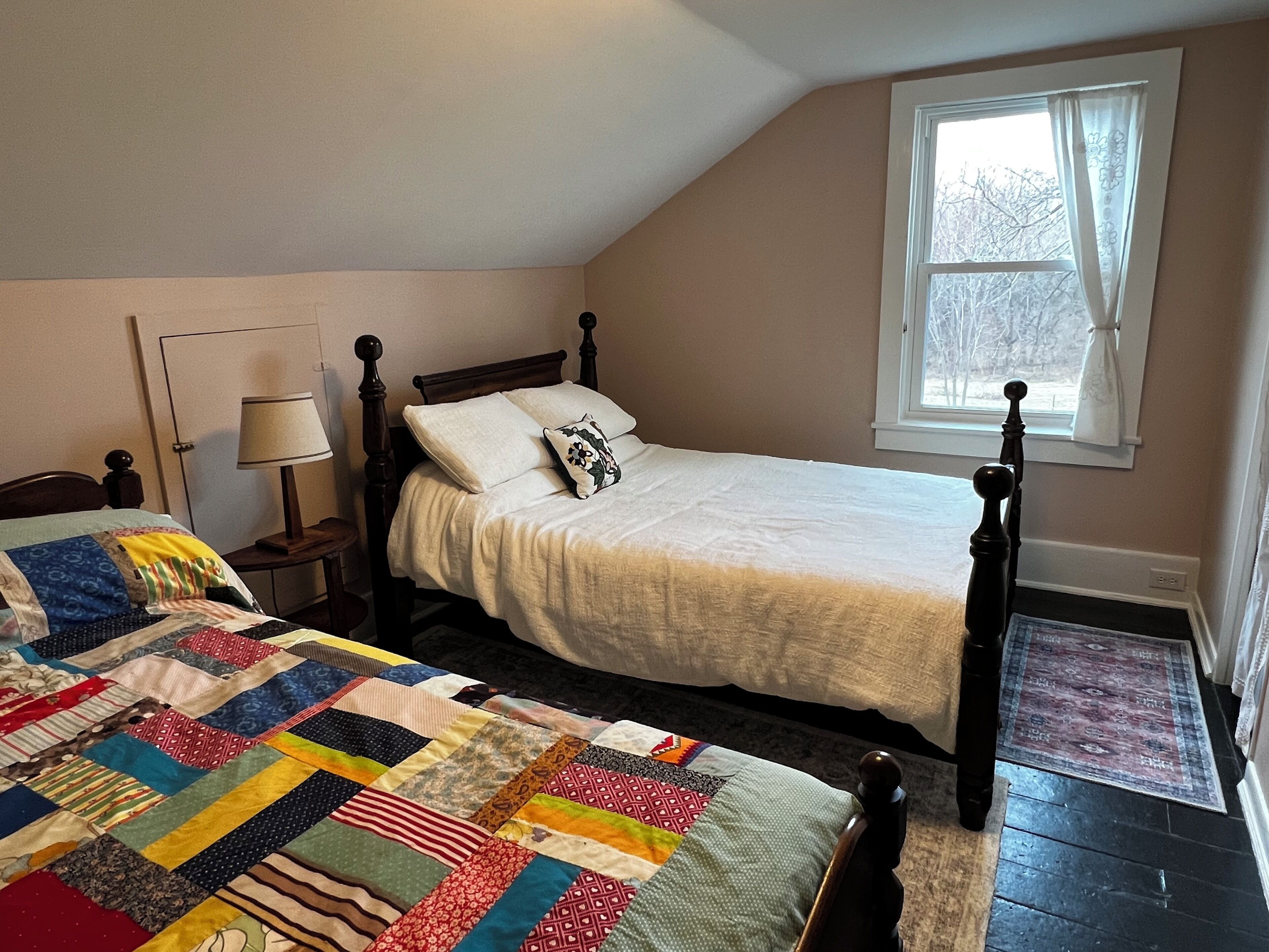 A bedroom with two beds, a window with curtains, a lamp, and a patchwork quilt on one bed.