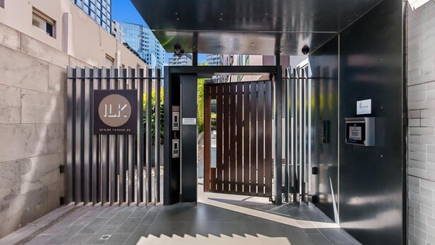 Une entrée moderne avec une grille métallique, une enseigne portant les initiales « JLX » et une adresse, ainsi qu’une caméra de surveillance.