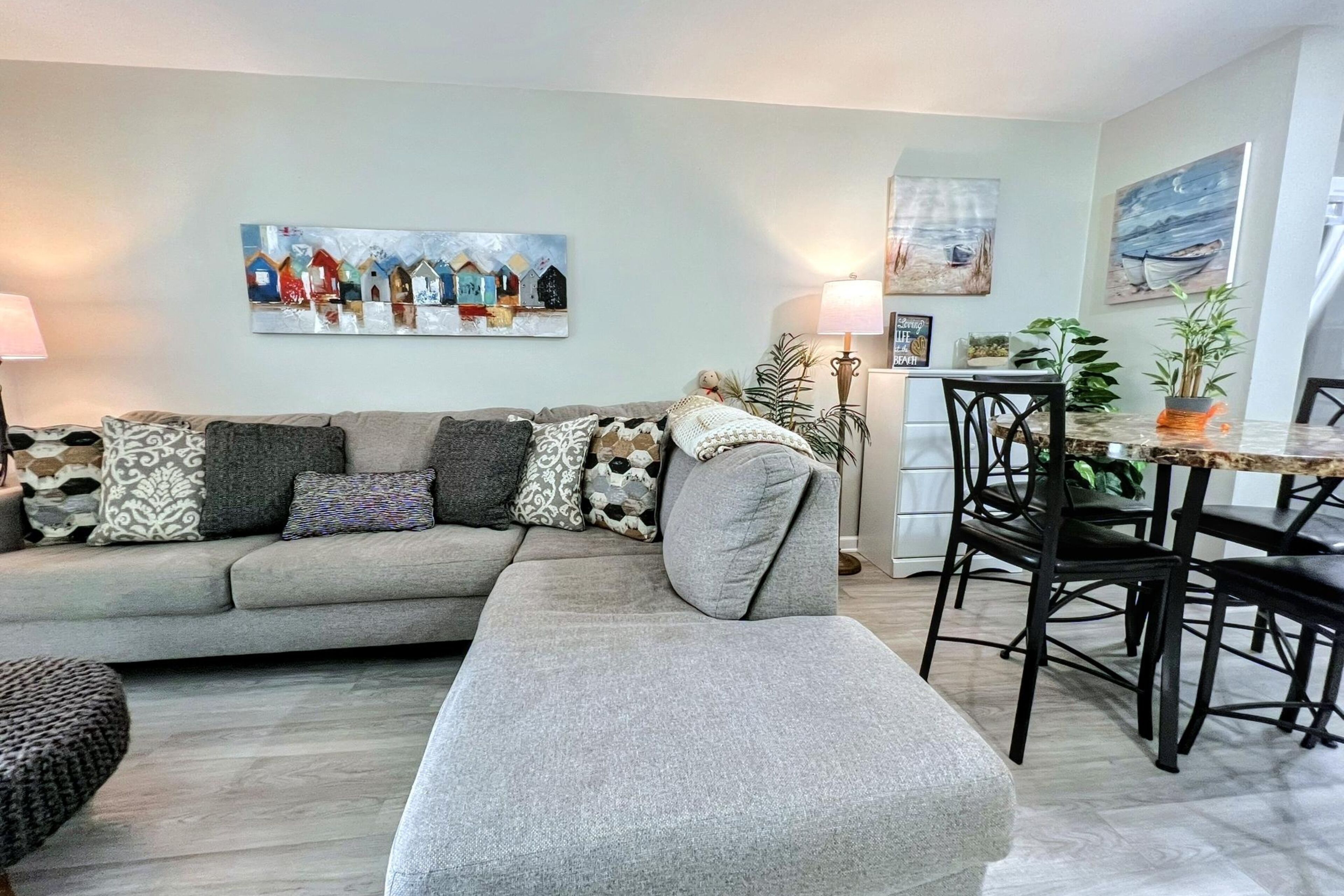 Een woonkamer met een grijze bank, gestreepte kussens, een eettafel met stoelen, en wanddecoratie waarop huizen en een boot te zien zijn.