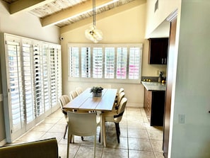 A dining area with a wooden table and chairs, large windows with blinds, and a kitchen visible through the window.