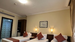A hotel room with three single beds, each with a patterned bedspread and matching pillows. There is a framed Arabic calligraphy on the wall, a ceiling light, and a window with a checkered pattern. - ASMA MAKKAH TAYSEER (Makkah)