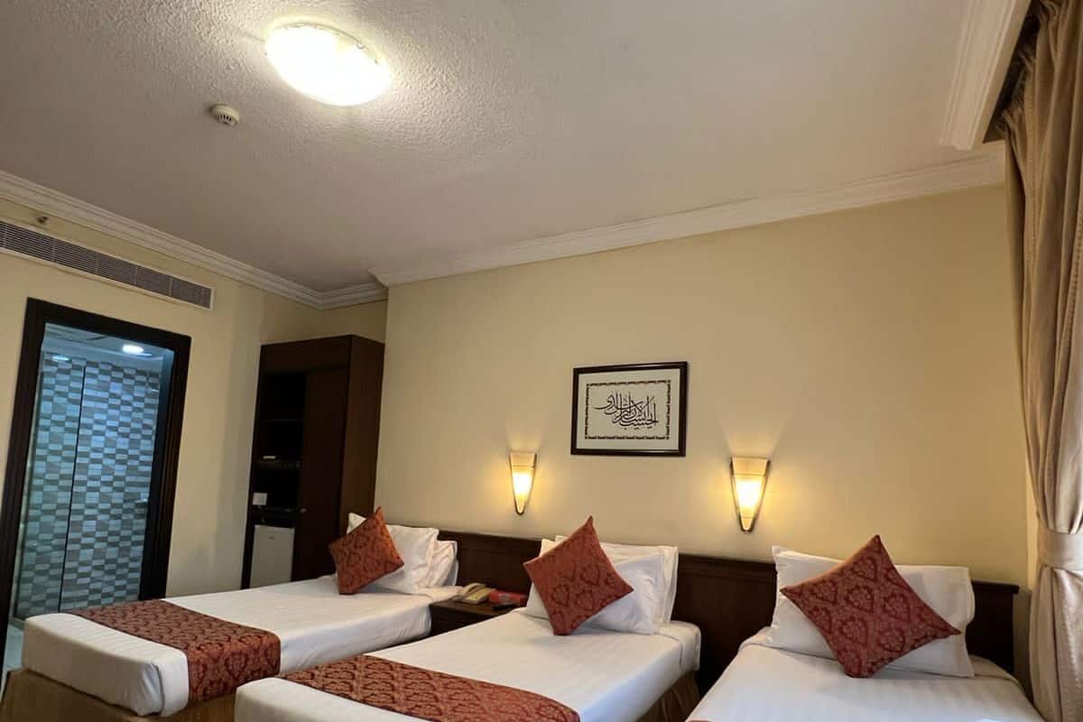 A hotel room with three single beds, each with a patterned bedspread and matching pillows. There is a framed Arabic calligraphy on the wall, a ceiling light, and a window with a checkered pattern.