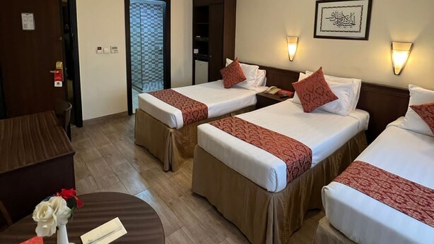A hotel room with three beds, a small table with a vase and a card, and a door leading to another room. - ASMA MAKKAH TAYSEER (Makkah)