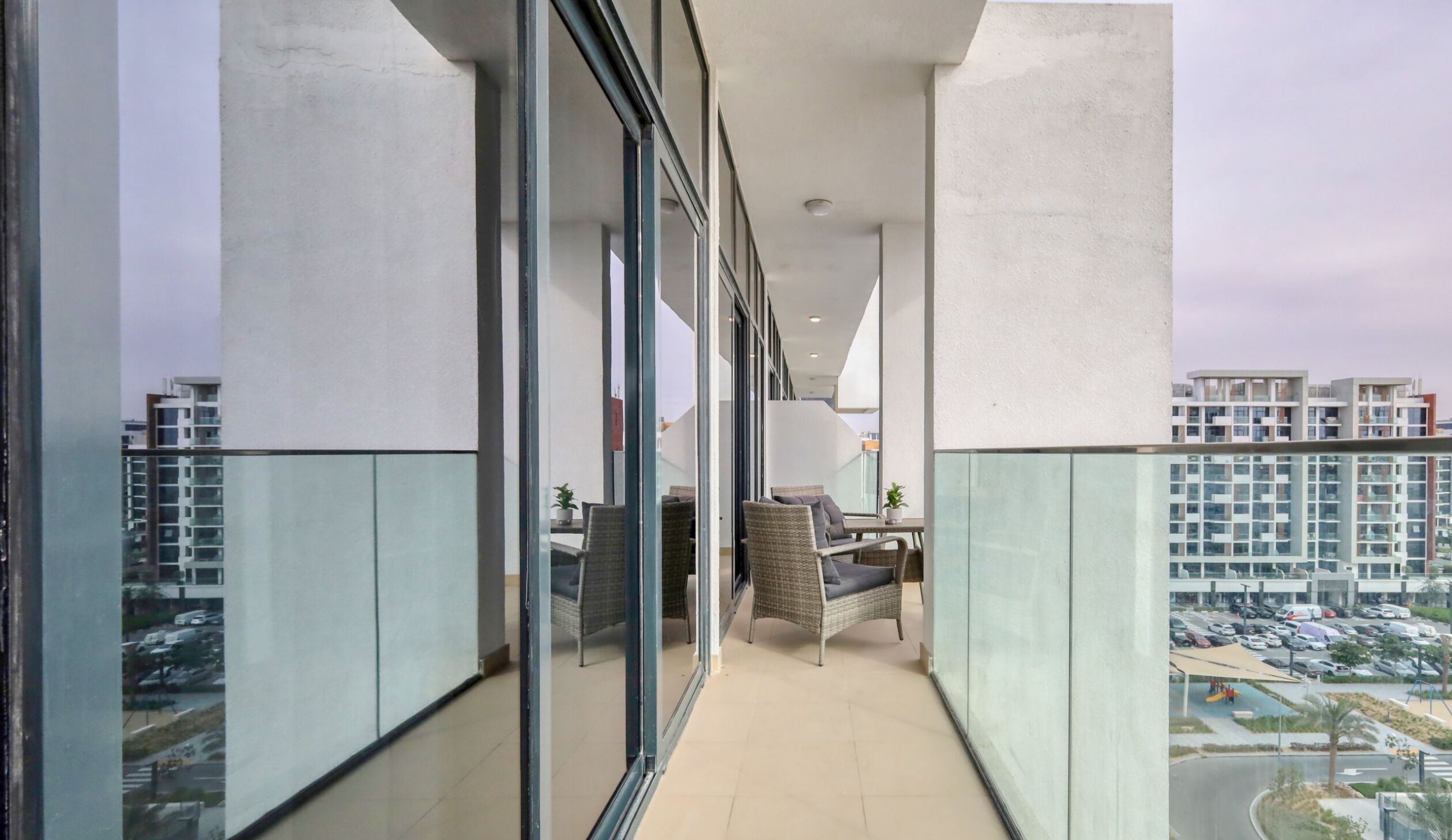 A modern balcony with glass walls, wicker furniture, and a view of other buildings and a parking area.