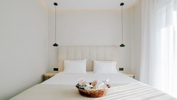 A neatly made bed with a white comforter, a wicker basket with bottles and snacks, and two bedside tables with lamps. - Aelia Prime Experience 3 (Kos)