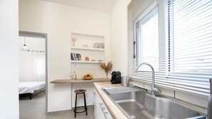 A compact kitchen with a sink, countertop, and a small stool. There is a window with blinds and a shelf with decorative items. - Coastal Elegance Suite 1Min to the Beach (Kos)