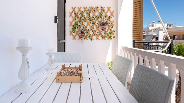A white wooden table with a box of snacks, two white candle holders, and a lattice wall with hanging planters on a balcony. - Beachside Retreat Steps to Kos (Kos)