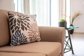 A beige sofa with a patterned pillow, a glass table with a plant, and a potted plant in the background.