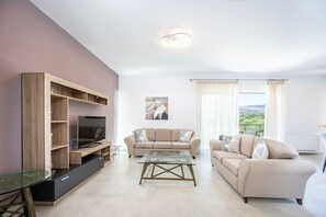 A modern living room with a TV on a wooden entertainment unit, two beige sofas, a glass coffee table, and a view of greenery through large windows.