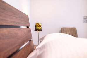 Une chambre d’hôtel avec une tête de lit en bois, un lit avec une literie blanche, une lampe avec un abat-jour doré et un fauteuil beige.