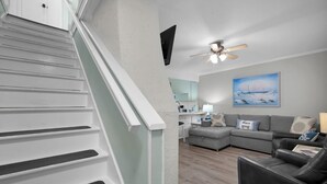 Una escalera con barandillas blancas, un sofá seccional gris, un sillón de cuero negro y un ventilador de techo.