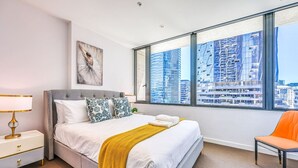 Habitación de hotel con una cama grande, una mesita de noche con lámpara, una silla y vistas a edificios altos.