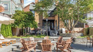 An outdoor patio with Adirondack chairs, a stone fire pit, and a view of a brick building with a stone facade.