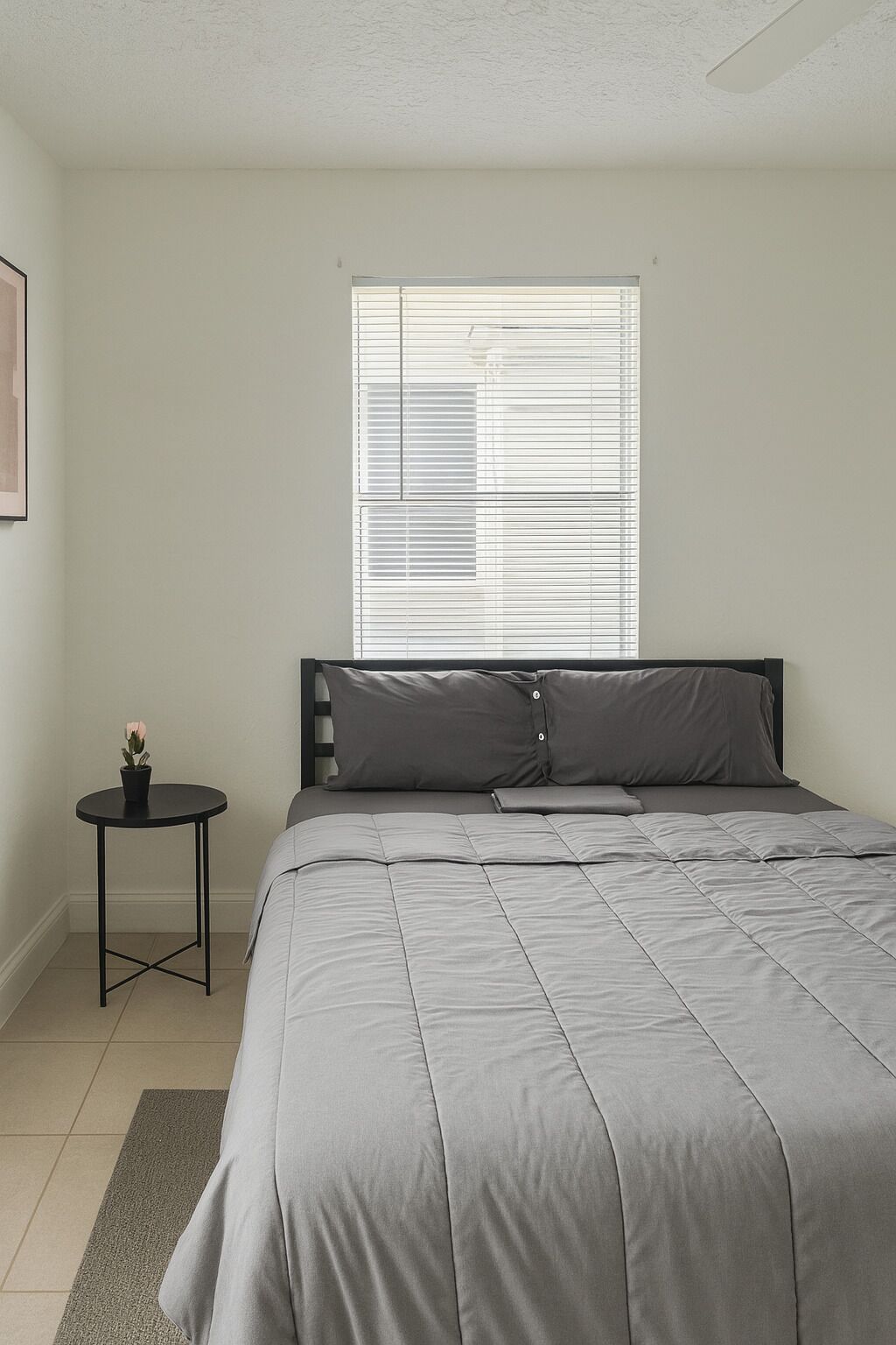 A bedroom with a bed, a small side table, a potted plant, and a window with blinds.