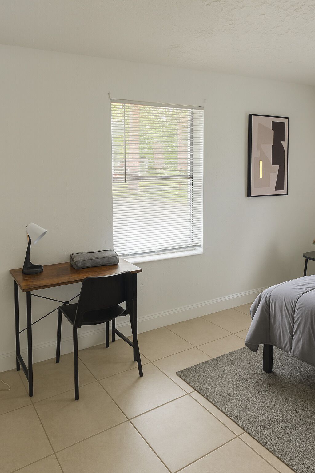 A bedroom with a bed, a desk with a chair, a window with blinds, and a framed picture on the wall.