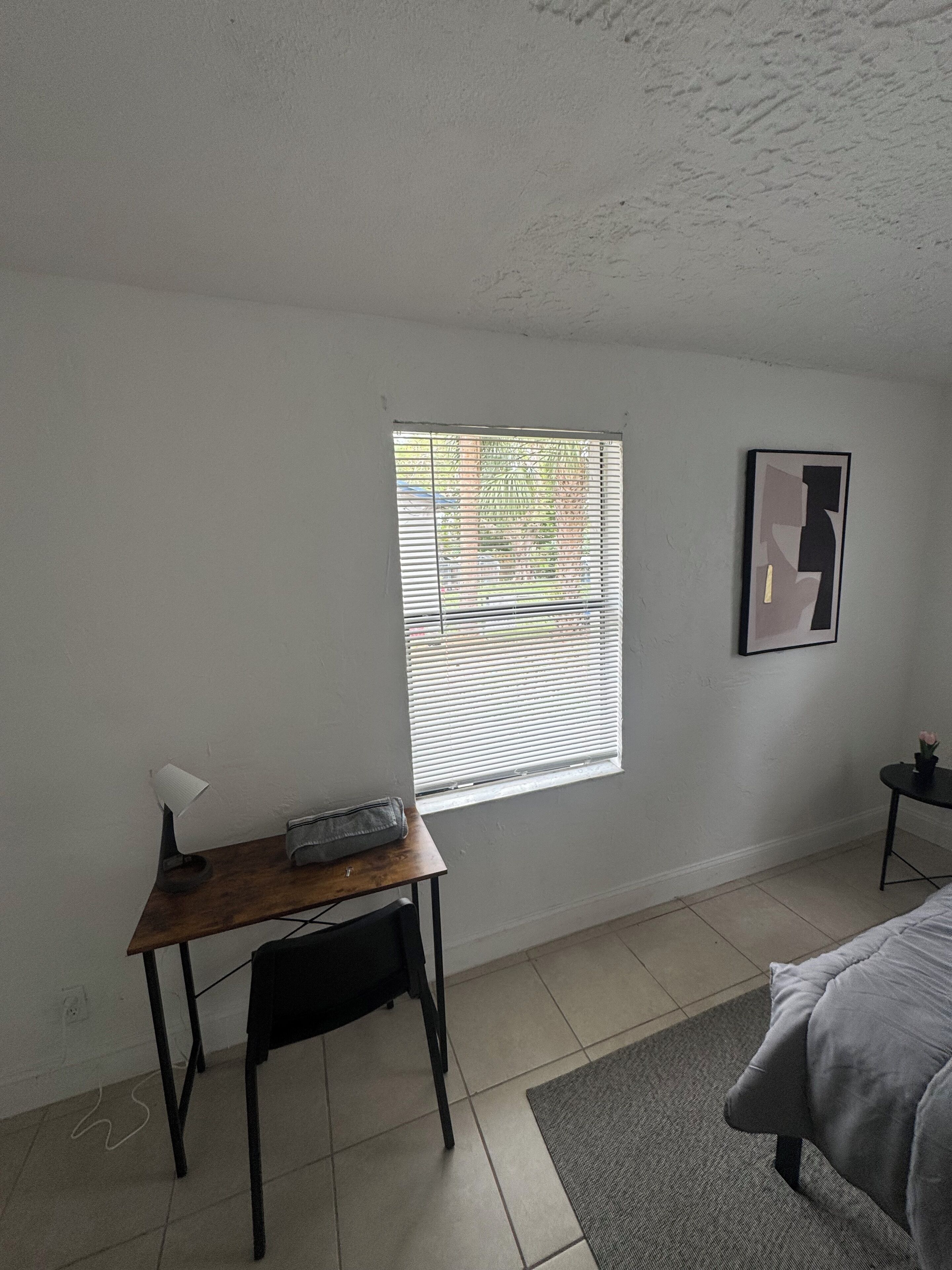 Une pièce comprenant un lit, un bureau avec une lampe, une fenêtre avec des stores et un tableau encadré au mur.