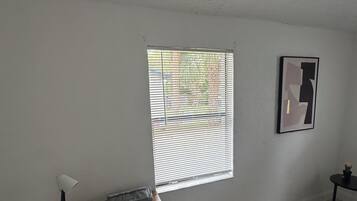 Une pièce comprenant un lit, un bureau avec une lampe, une fenêtre avec des stores et un tableau encadré au mur.