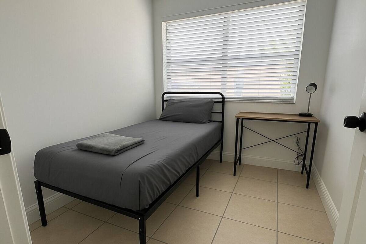 Een compacte, netjes ingerichte slaapkamer met een enkel bed, een bureau en een raam met zonwering.