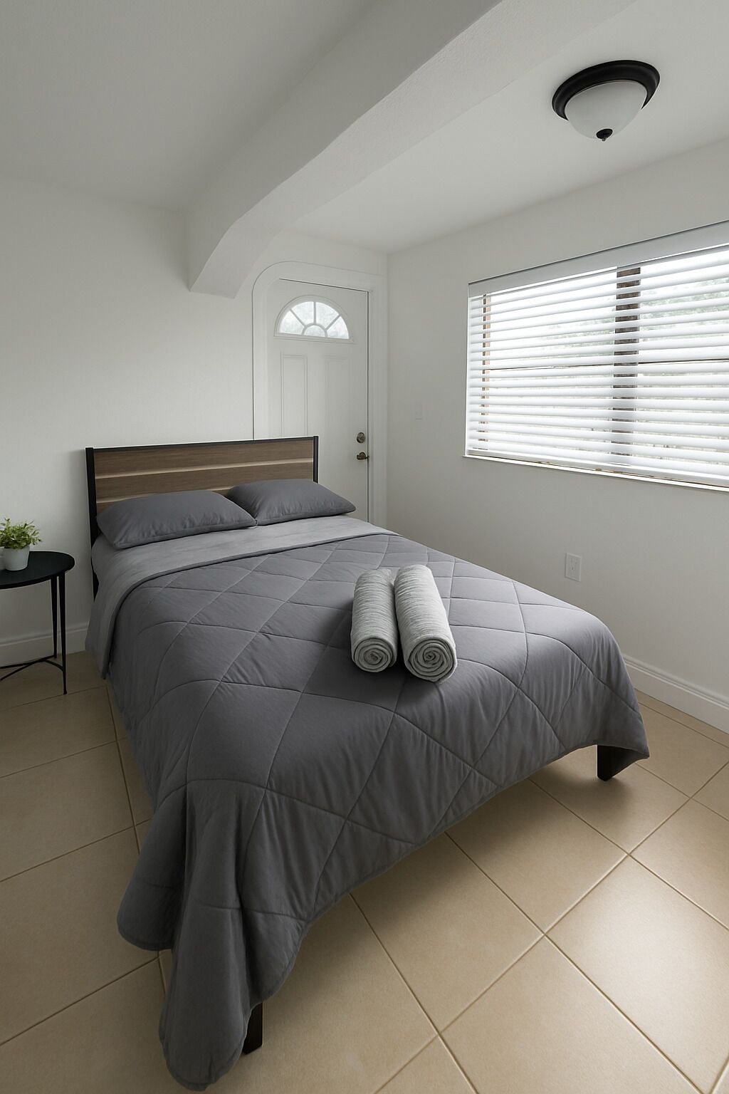 A bedroom with a bed, a nightstand, a potted plant, a window with blinds, and a door with a glass panel.
