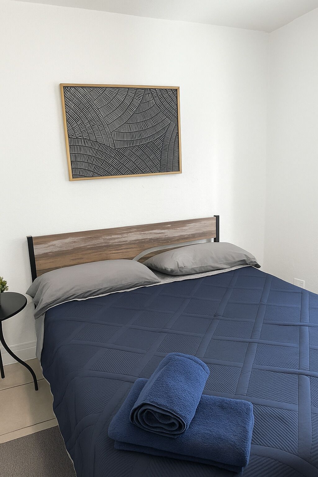 A neatly made bed with a blue quilt, two pillows, and a rolled blue towel on the side.