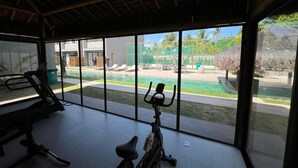 Ein Fitnessraum mit Trainingsgeräten, einem Schwimmbad und Blick auf eine tropische Außenanlage.